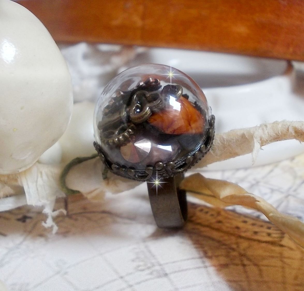 Bague Majesté Steampunk aux rouages mécaniques à laquelle s’ajoute une clé et un strass de couleur Siam et un strass en verre Doré. Le tout est mis sous un globe en verre sur une bague réglable laiton