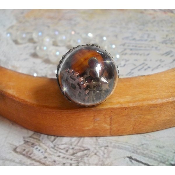 Bague Majest&eacute; Steampunk aux rouages m&eacute;caniques &agrave; laquelle s&rsquo;ajoute une cl&eacute; et un strass de couleur Siam et un strass en verre Dor&eacute;. Le tout est mis sous un globe en verre sur une bague r&eacute;glable laiton